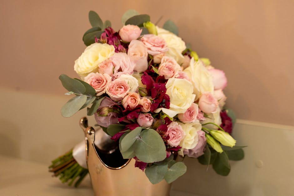 A floral bouquet featuring soft pink and white roses, complemented by deep burgundy alstroemeria and sprigs of eucalyptus with rounded, silvery-green leaves. The arrangement is styled in a rounded, lu