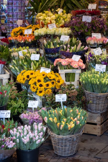 A vibrant display of fresh flowers at Spitalfields Florist, featuring various spring blooms arranged in baskets and tubs. Prominent in the foreground are yellow sunflowers and pale pink tulips with cl