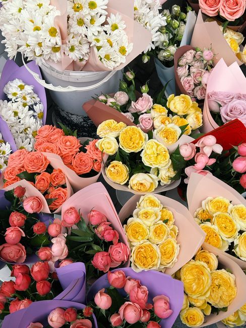 A vibrant display of fresh flower bouquets arranged in various pastel and bright wrapping papers, including pink, purple, and peach tissue wraps. The arrangements feature a mix of roses in shades of p