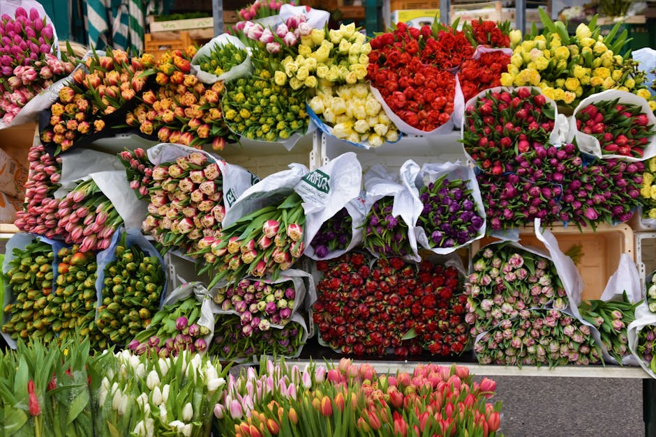 A vibrant display of fresh flower bouquets arranged in a florist shop, featuring tulips, roses, and other blooms in shades of pink, red, white, purple, and yellow. The flowers are wrapped in white and