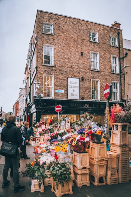 An outdoor flower market scene in Spitalfields featuring a busy sidewalk filled with vibrant floral bouquets arranged on wooden crates and tables. The display includes a variety of fresh flowers such 