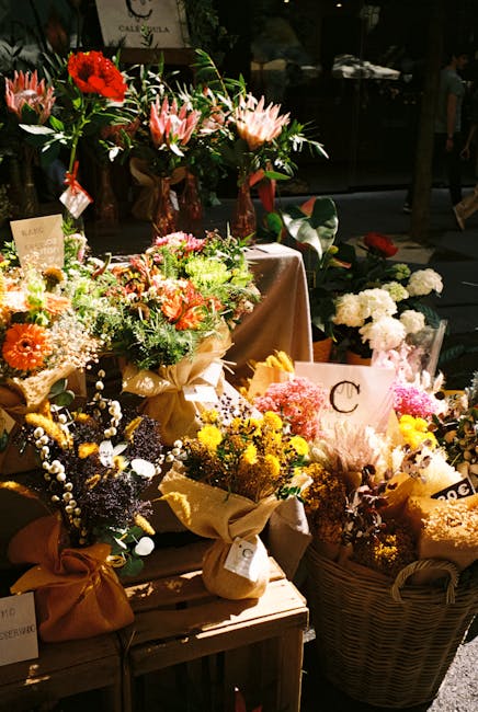 A display of various floral arrangements and bouquets situated outdoors at the Spitalfields Market, featuring an assortment of fresh flowers in vibrant colors including reds, pinks, peaches, yellows, 